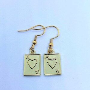 May include: A pair of gold-colored earrings featuring the Ace of Hearts playing card design. Each earring has a rectangular pendant with a heart symbol and the letter "A" at the top, with a small "V" at the bottom.
