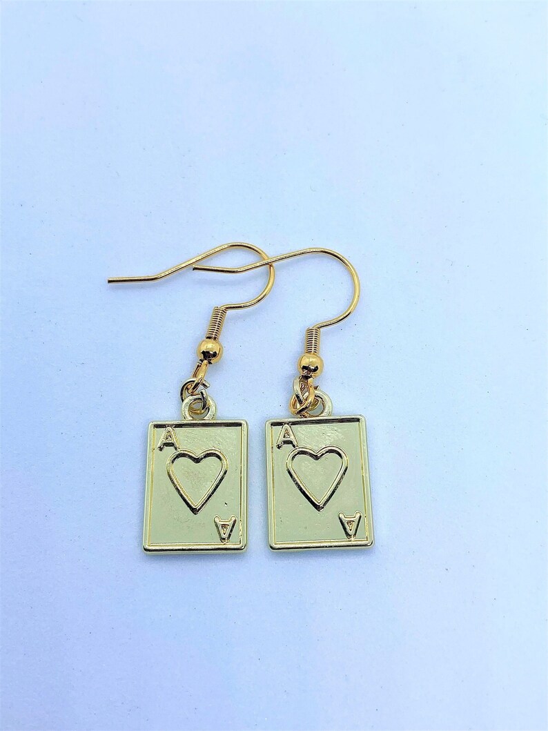 May include: A pair of gold-colored earrings featuring the Ace of Hearts playing card design. Each earring has a rectangular pendant with a heart symbol and the letter "A" at the top, with a small "V" at the bottom.