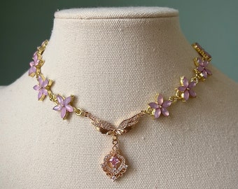 Puede incluir: Un collar de oro con un diseño de flores rosas y un colgante con un cristal en el centro. El colgante tiene forma de corazón estilizado con alas.