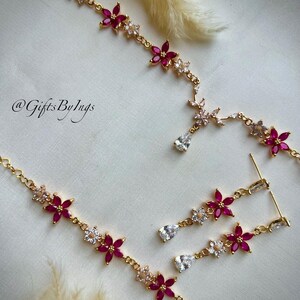 May include: A gold-toned necklace and earring set featuring alternating pink and clear floral-shaped stones. The necklace includes a central teardrop-shaped clear stone pendant. The text @GiftsByIngs is visible on the left side of the image.