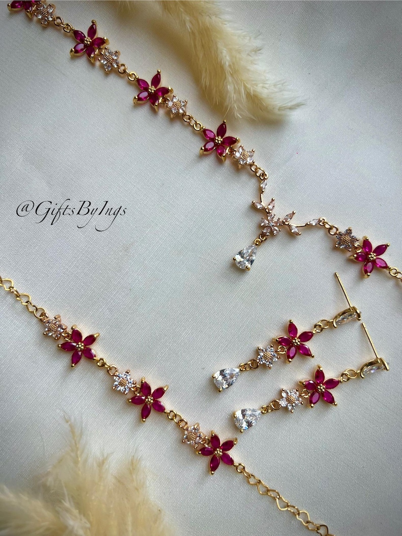 May include: A gold-toned necklace and earring set featuring alternating pink and clear floral-shaped stones. The necklace includes a central teardrop-shaped clear stone pendant. The text @GiftsByIngs is visible on the left side of the image.