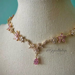 May include: A gold-tone necklace featuring a series of pink and clear crystal flower-shaped links connected by a delicate chain. The center of the necklace displays a floral motif with a dangling pear-shaped pink crystal pendant. Text reads @GiftsByIngs.