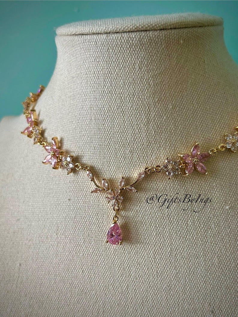 May include: A gold-tone necklace featuring a series of pink and clear crystal flower-shaped links connected by a delicate chain. The center of the necklace displays a floral motif with a dangling pear-shaped pink crystal pendant. Text reads @GiftsByIngs.