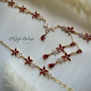 May include: A gold-toned jewelry set featuring a necklace and matching drop earrings. The pieces are adorned with red, petal-shaped stones and small, clear, sparkling stones arranged in flower patterns. The text @GiftsByIngs is visible.