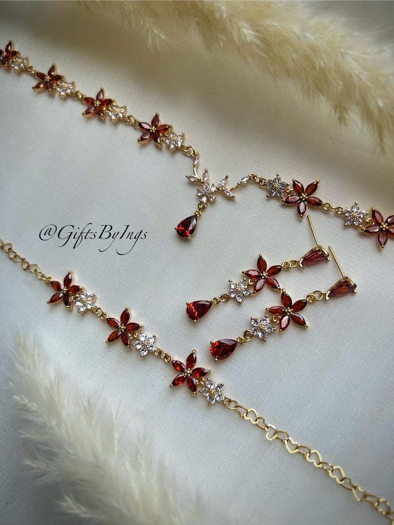 May include: A gold-toned jewelry set featuring a necklace and matching drop earrings. The pieces are adorned with red, petal-shaped stones and small, clear, sparkling stones arranged in flower patterns. The text @GiftsByIngs is visible.