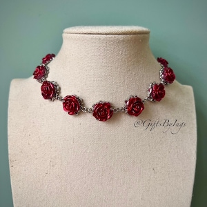 Può includere: Collana con pendenti a forma di rosa rossa, esposta su un manichino. Le rose, collegate da una catena argentata, hanno una finitura lucida. Il testo "@GiftsByIngs" è visibile. È un gioiello decorativo.