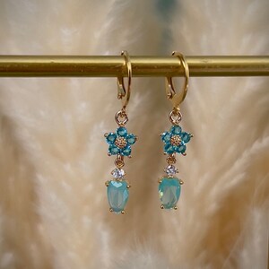 Puede incluir: Pendientes de aro dorados con un dise&ntilde;o de flor azul y una piedra azul claro en forma de l&aacute;grima.