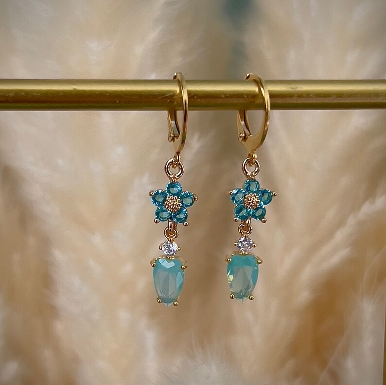 Puede incluir: Pendientes de aro dorados con un dise&ntilde;o de flor azul y una piedra azul claro en forma de l&aacute;grima.