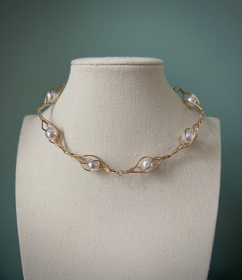 May include: A gold wire necklace with white pearls set in delicate wire cages. The necklace is displayed on a white mannequin.