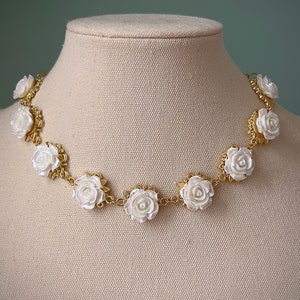 May include: A gold chain necklace with white porcelain rose accents. The necklace is displayed on a fabric-covered mannequin.