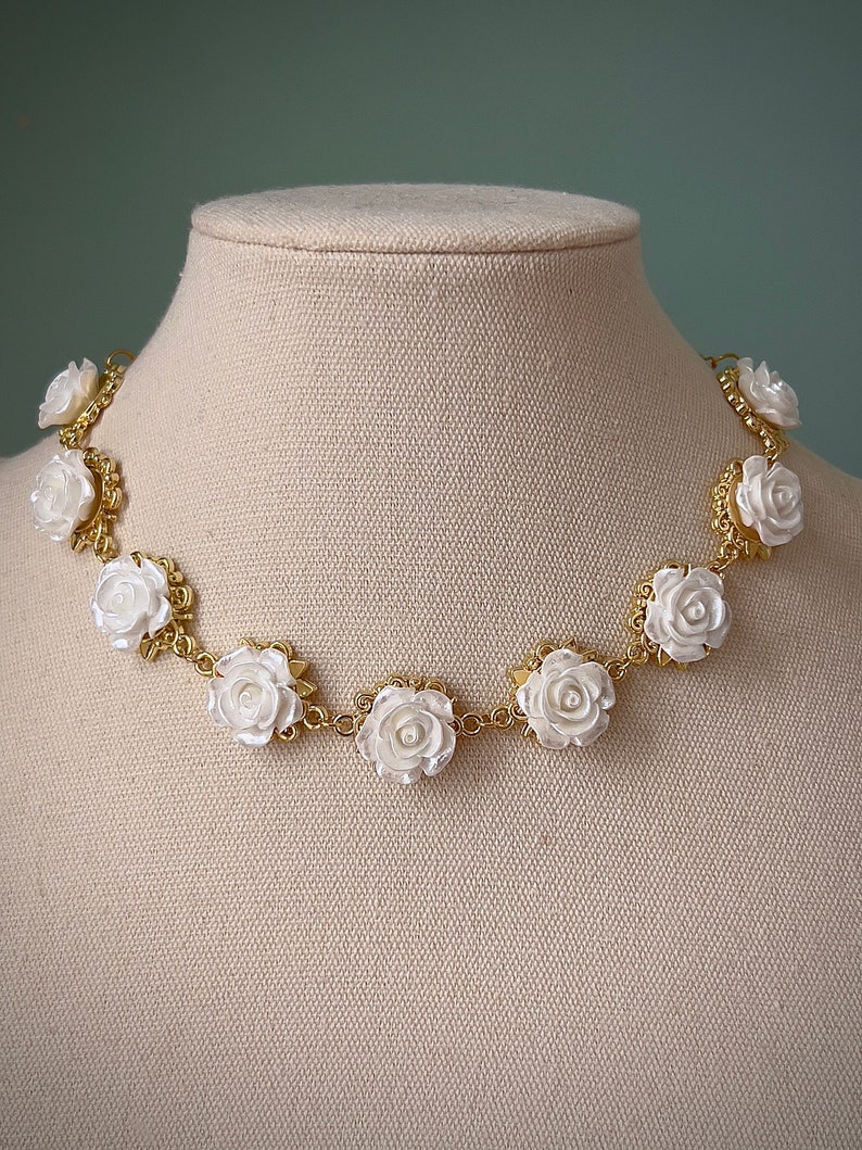 May include: A gold chain necklace with white porcelain rose accents. The necklace is displayed on a fabric-covered mannequin.