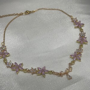 May include: A gold necklace with a delicate chain and pink flower accents. The flowers are set with clear rhinestones.