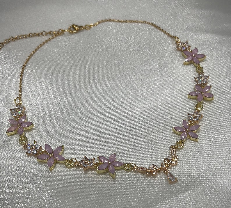 May include: A gold necklace with a delicate chain and pink flower accents. The flowers are set with clear rhinestones.