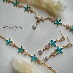 May include: A gold-toned jewelry set featuring a necklace and matching drop earrings. The pieces are adorned with alternating turquoise and clear floral-shaped crystals, with a single pear-shaped clear crystal drop. The text @GiftsByIngs is visible.