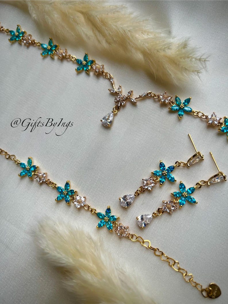 May include: A gold-toned jewelry set featuring a necklace and matching drop earrings. The pieces are adorned with alternating turquoise and clear floral-shaped crystals, with a single pear-shaped clear crystal drop. The text @GiftsByIngs is visible.