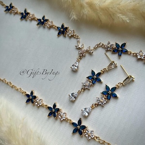 May include: A gold-toned jewelry set featuring a necklace and matching drop earrings. The pieces are adorned with blue marquise-cut stones arranged in flower shapes, interspersed with clear crystals. The text @GiftsByIngs is visible.