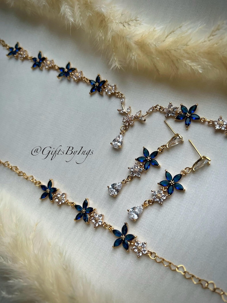 May include: A gold-toned jewelry set featuring a necklace and matching drop earrings. The pieces are adorned with blue marquise-cut stones arranged in flower shapes, interspersed with clear crystals. The text @GiftsByIngs is visible.