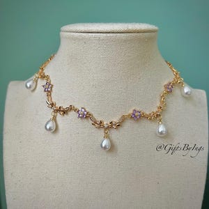 May include: A gold-toned necklace with a floral design, featuring purple flower accents and dangling pear-shaped pearls. The necklace is displayed on a mannequin, showcasing its delicate details and elegant style. The necklace is a beautiful piece of jewelry.