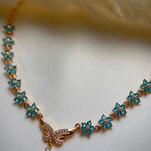 Puede incluir: Un collar de oro con un colgante de mariposa y detalles de flores azules. El colgante tiene una piedra de &oacute;palo en forma de l&aacute;grima.