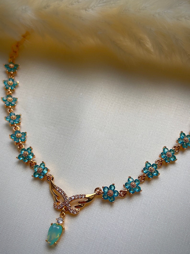 Puede incluir: Un collar de oro con un colgante de mariposa y detalles de flores azules. El colgante tiene una piedra de &oacute;palo en forma de l&aacute;grima.