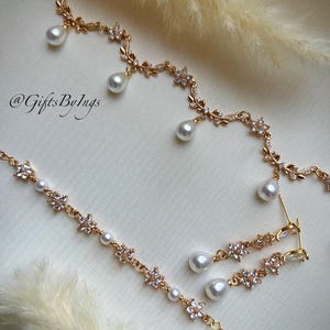 May include: Gold-toned jewelry set featuring a necklace, bracelet, and earrings. The pieces are adorned with floral and leaf designs, accented with clear crystals and dangling white pearls. The image includes the text "@GiftsByIngs".