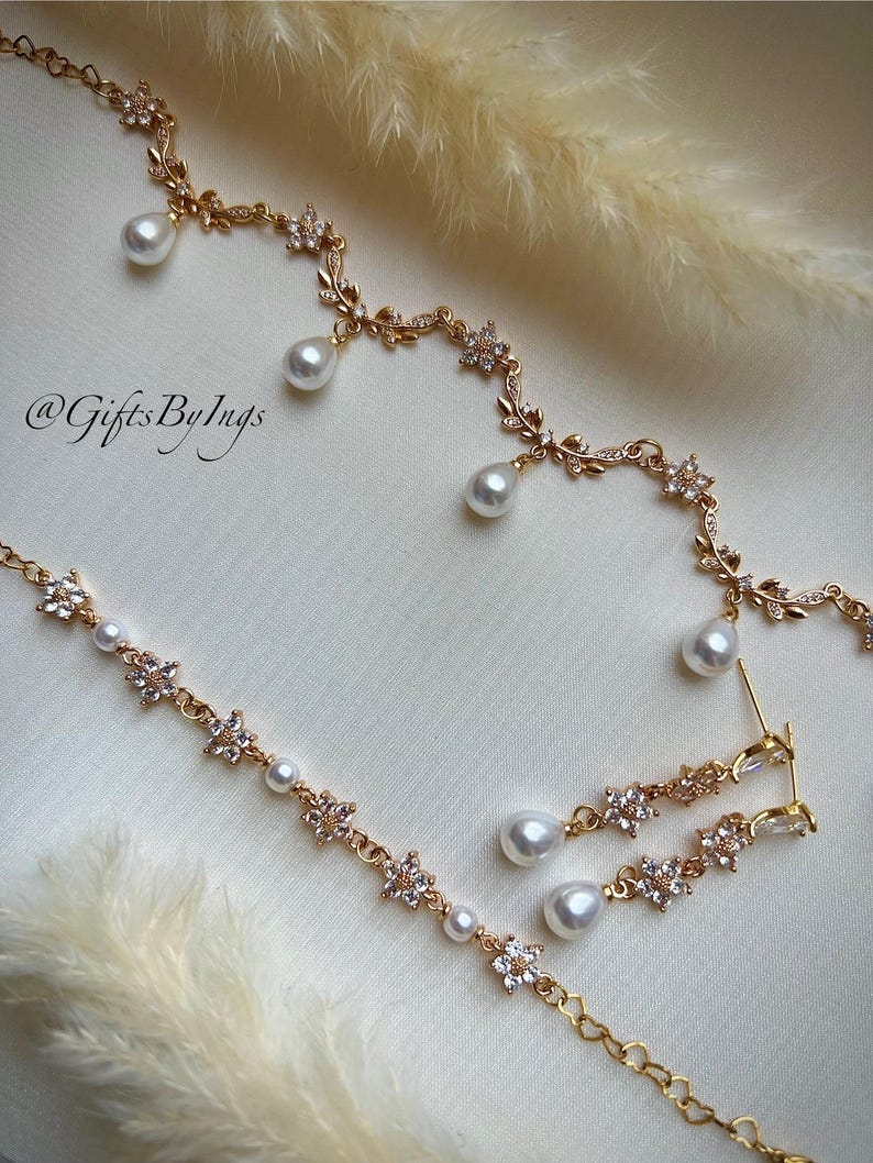 May include: Gold-toned jewelry set featuring a necklace, bracelet, and earrings. The pieces are adorned with floral and leaf designs, accented with clear crystals and dangling white pearls. The image includes the text "@GiftsByIngs".