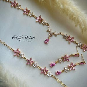 May include: A gold-toned jewelry set featuring a necklace and matching drop earrings. The pieces are adorned with pink and clear floral-shaped crystals and teardrop-shaped pink stones. The text @GiftsByIngs is visible on the surface.