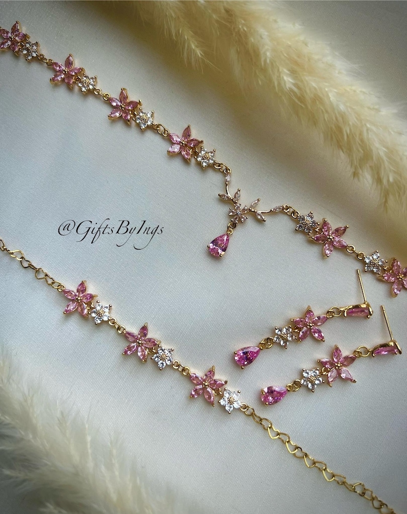 May include: A gold-toned jewelry set featuring a necklace and matching drop earrings. The pieces are adorned with pink and clear floral-shaped crystals and teardrop-shaped pink stones. The text @GiftsByIngs is visible on the surface.