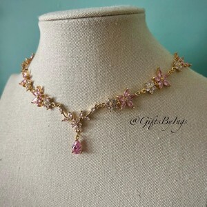 May include: A gold-tone necklace featuring a series of pink and clear crystal flower-shaped links connected by thin metal chains, with a central teardrop-shaped pink crystal pendant. The text @GiftsByIngs is visible on the right side.