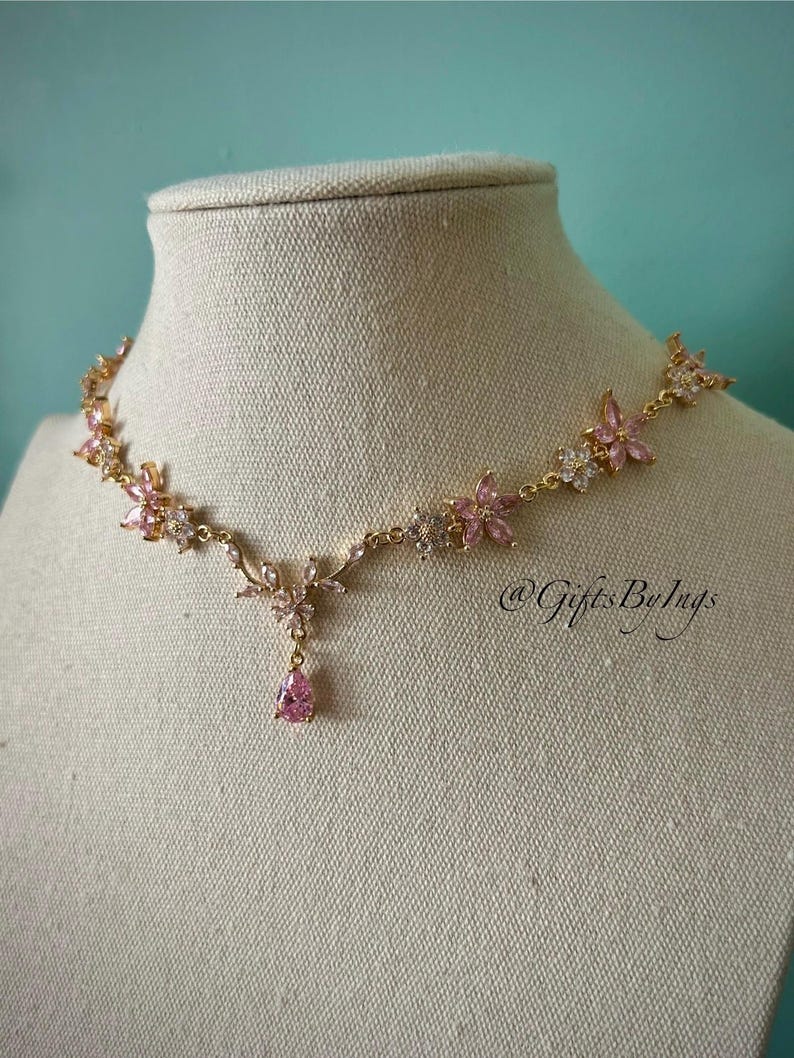 May include: A gold-tone necklace featuring a series of pink and clear crystal flower-shaped links connected by thin metal chains, with a central teardrop-shaped pink crystal pendant. The text @GiftsByIngs is visible on the right side.
