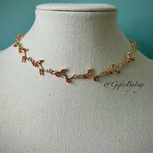 May include: A gold necklace with a delicate vine design and small red gemstones. The necklace is on a white mannequin bust.