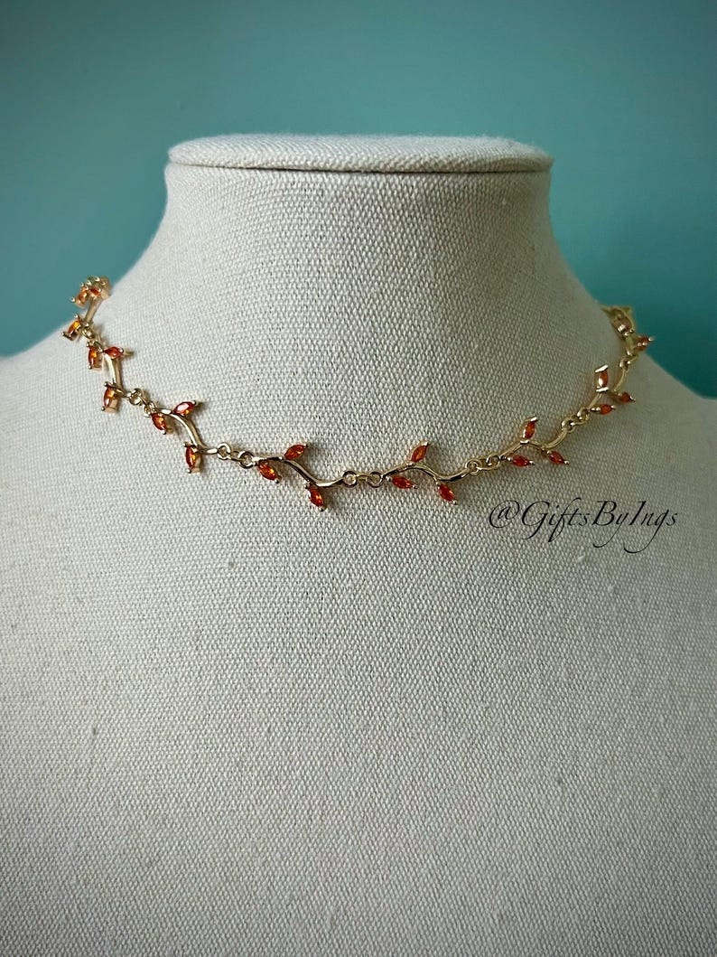 May include: A gold necklace with a delicate vine design and small red gemstones. The necklace is on a white mannequin bust.