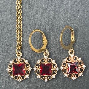Pode incluir: Um conjunto de colar e brincos de ouro com uma pedra preciosa quadrada vermelha cercada por um design de filigrana. O colar é de estilo corrente e os brincos são de estilo pendente.