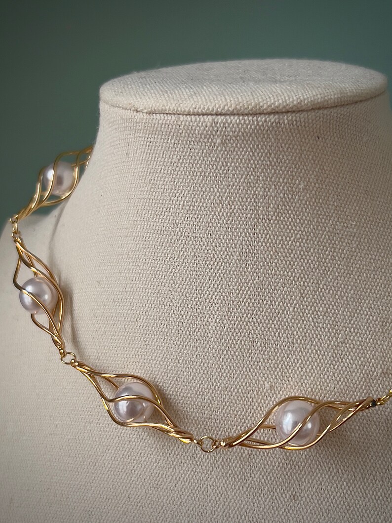 May include: A gold necklace with a delicate wire design that holds white pearls. The necklace is displayed on a white fabric mannequin.