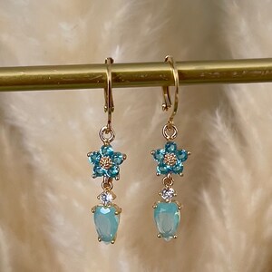 Puede incluir: Un par de pendientes colgantes de oro con una flor azul y detalles de cristal en forma de l&aacute;grima.