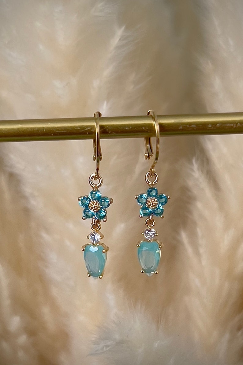 Puede incluir: Un par de pendientes colgantes de oro con una flor azul y detalles de cristal en forma de l&aacute;grima.