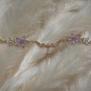 May include: A gold chain bracelet with a delicate floral design. The flowers are made of light purple enamel and have clear rhinestones in the center. The bracelet has a small pendant in the center with a clear rhinestone.