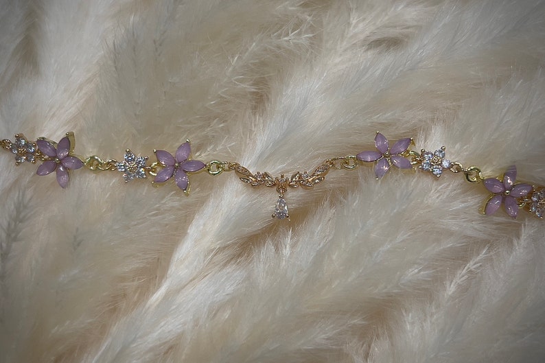 May include: A gold chain bracelet with a delicate floral design. The flowers are made of light purple enamel and have clear rhinestones in the center. The bracelet has a small pendant in the center with a clear rhinestone.