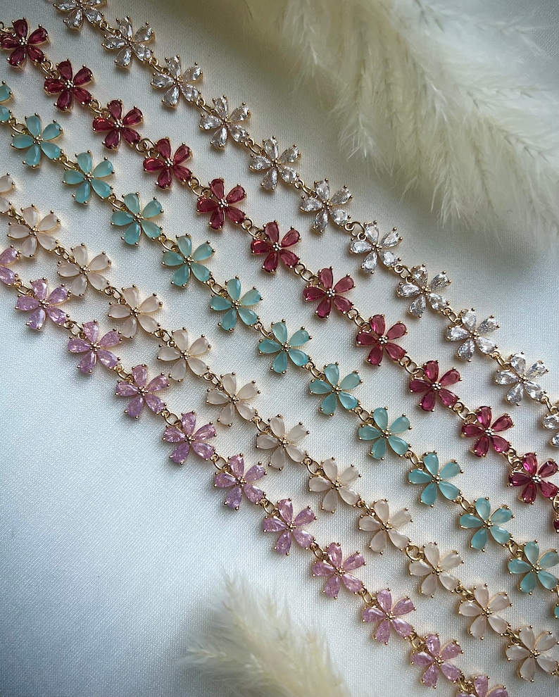 May include: A set of six gold flower chain necklaces with different colored flower accents. The necklaces feature pink, light blue, cream, and white flower accents.