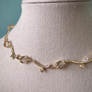 May include: A gold necklace with a delicate vine design featuring small leaves and clear stones. The necklace is displayed on a beige fabric mannequin.