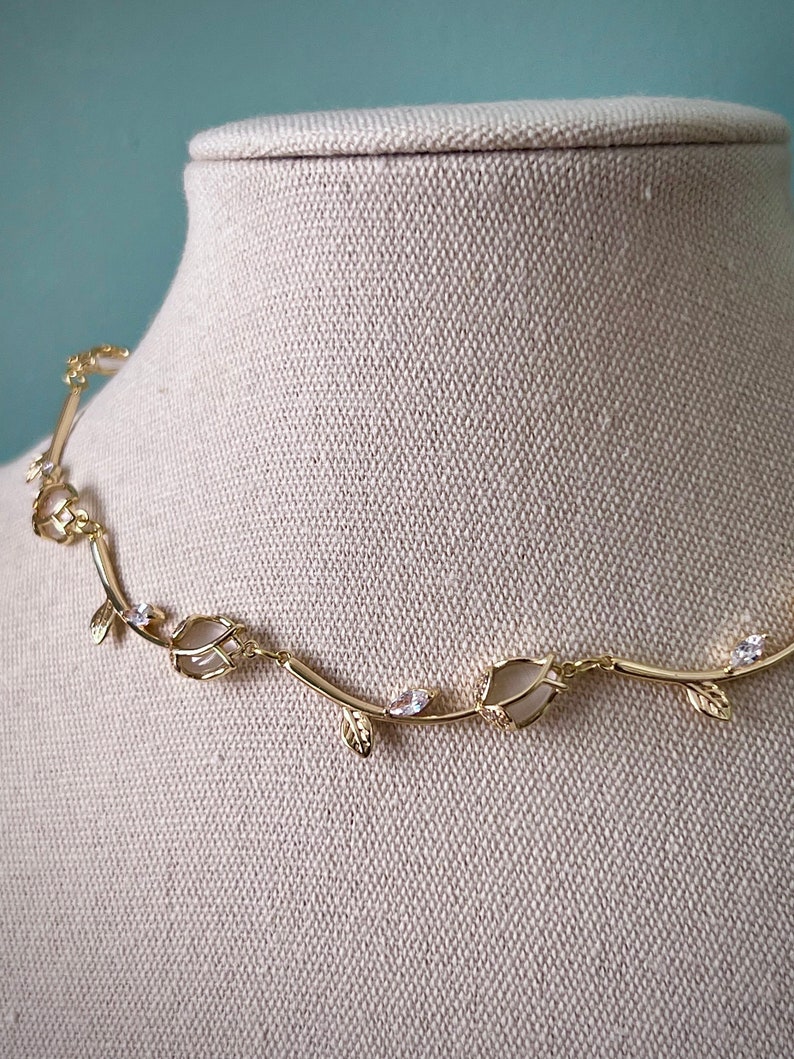 May include: A gold necklace with a delicate vine design featuring small leaves and clear stones. The necklace is displayed on a beige fabric mannequin.