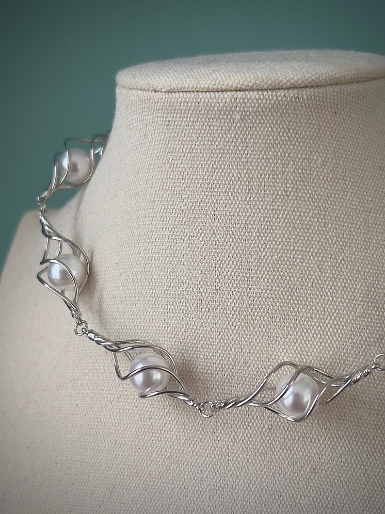 May include: A silver wire necklace with a delicate, twisted design. The necklace features five white pearls, each encased in a wire cage.