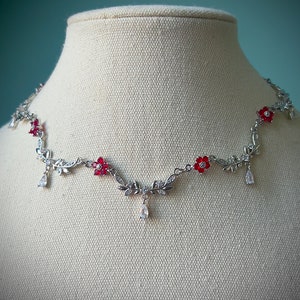 May include: Silver necklace with red flower accents and clear crystal drops. The necklace features a delicate vine design with leaves and flowers.