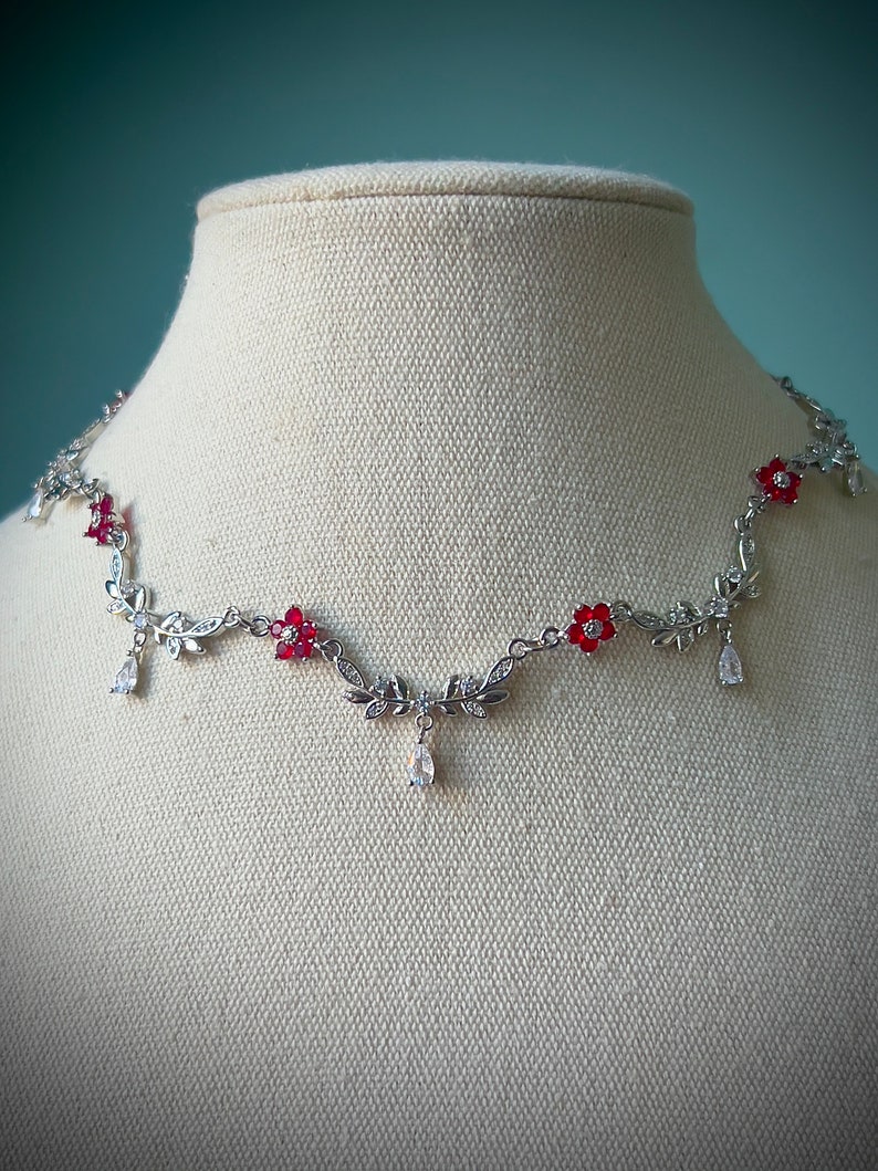 May include: Silver necklace with red flower accents and clear crystal drops. The necklace features a delicate vine design with leaves and flowers.