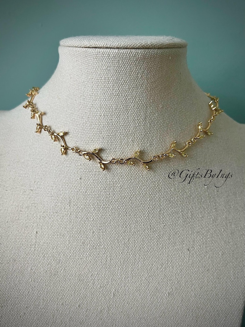 May include: A gold necklace with a delicate vine design and small clear stones. The necklace is on a white mannequin bust.