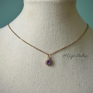 Puede incluir: Un collar dorado con un pequeño colgante ovalado de amatista púrpura, rodeado de piedras claras. El collar se exhibe en un maniquí sobre un fondo neutro. El texto "@GiftsByIngs" es visible. La cadena es delicada.