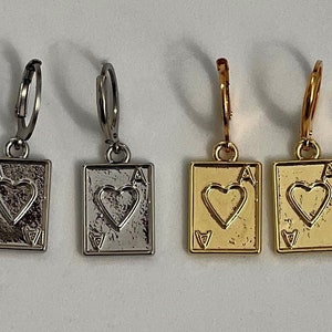 May include: Four earrings featuring a playing card design. Two are silver-toned, and two are gold-toned. Each earring depicts the Ace of Hearts card, with a heart symbol in the center and the letter "A" in the corners. The earrings have a hoop closure.