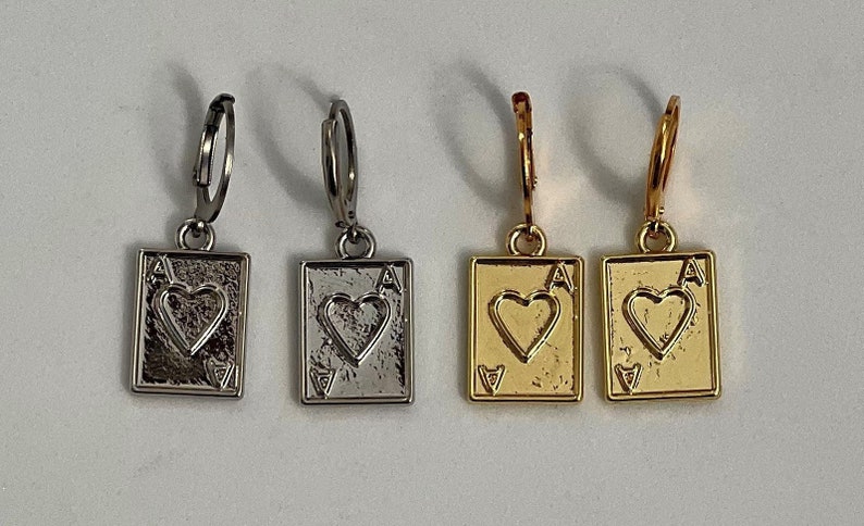May include: Four earrings featuring a playing card design. Two are silver-toned, and two are gold-toned. Each earring depicts the Ace of Hearts card, with a heart symbol in the center and the letter "A" in the corners. The earrings have a hoop closure.