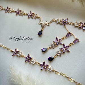 May include: A gold-toned jewelry set featuring a necklace and matching drop earrings. The pieces are adorned with purple and clear floral-shaped crystals and dark purple teardrop-shaped stones. The text @GiftsByIngs is visible in the center.