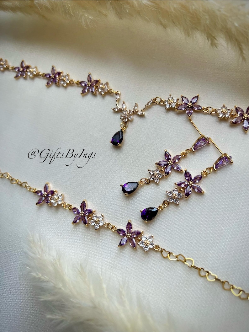 May include: A gold-toned jewelry set featuring a necklace and matching drop earrings. The pieces are adorned with purple and clear floral-shaped crystals and dark purple teardrop-shaped stones. The text @GiftsByIngs is visible in the center.